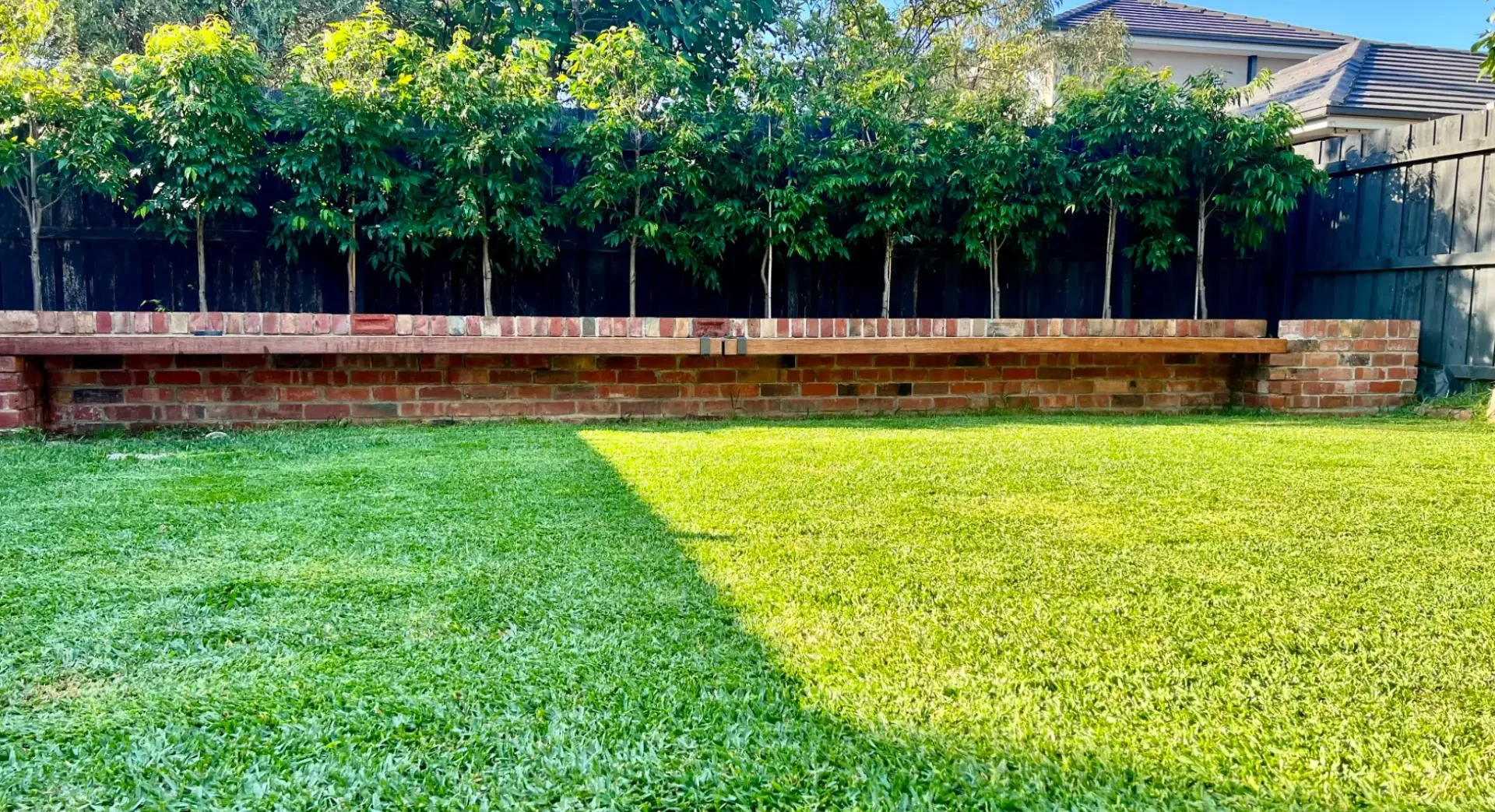 A brick bench with wooden seat in a backyard garden with green grass and trees.